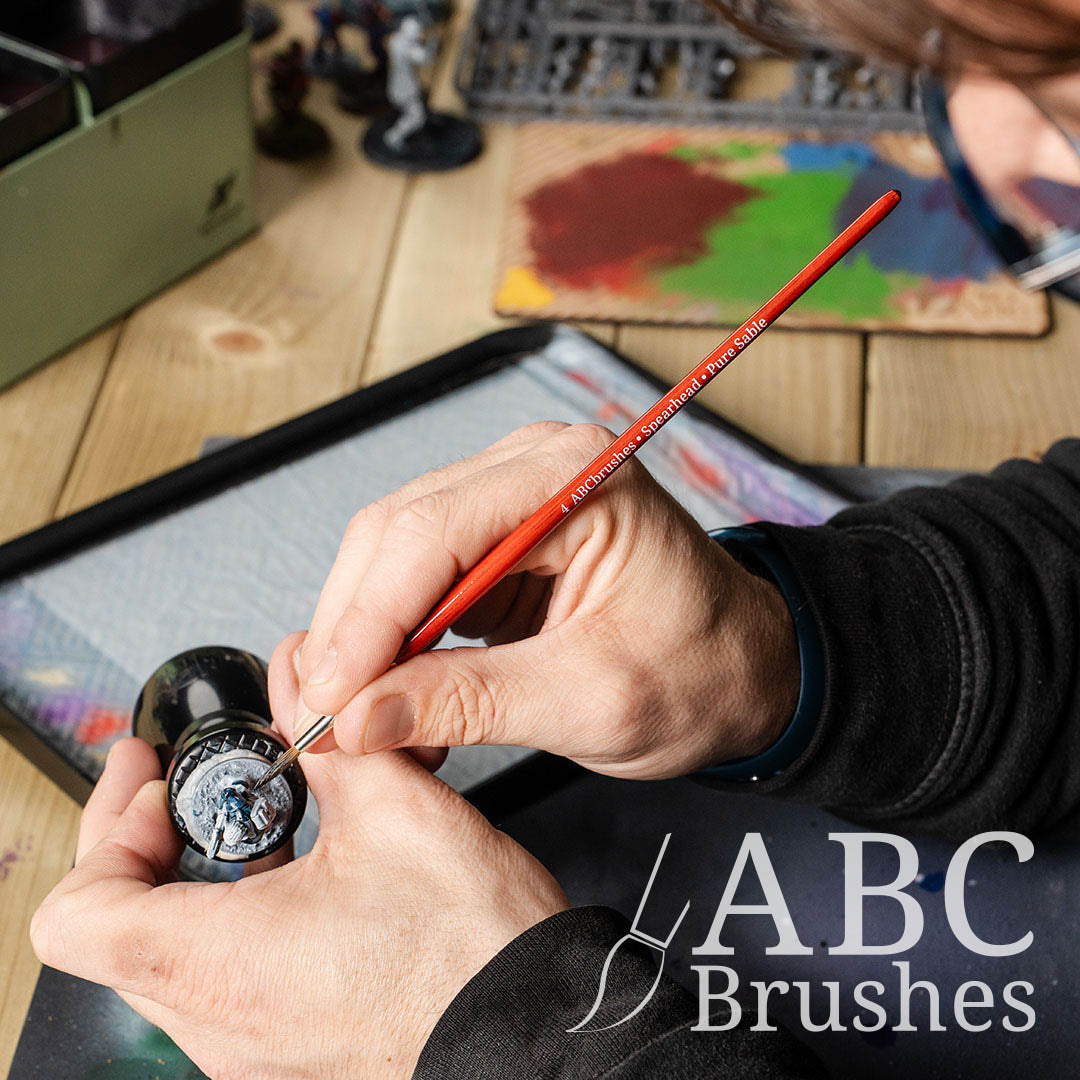 Painting blue detailing onto a mini with a ABC Brushes Spearhead paint brush, with a wet palette and dry brush board in the background. 