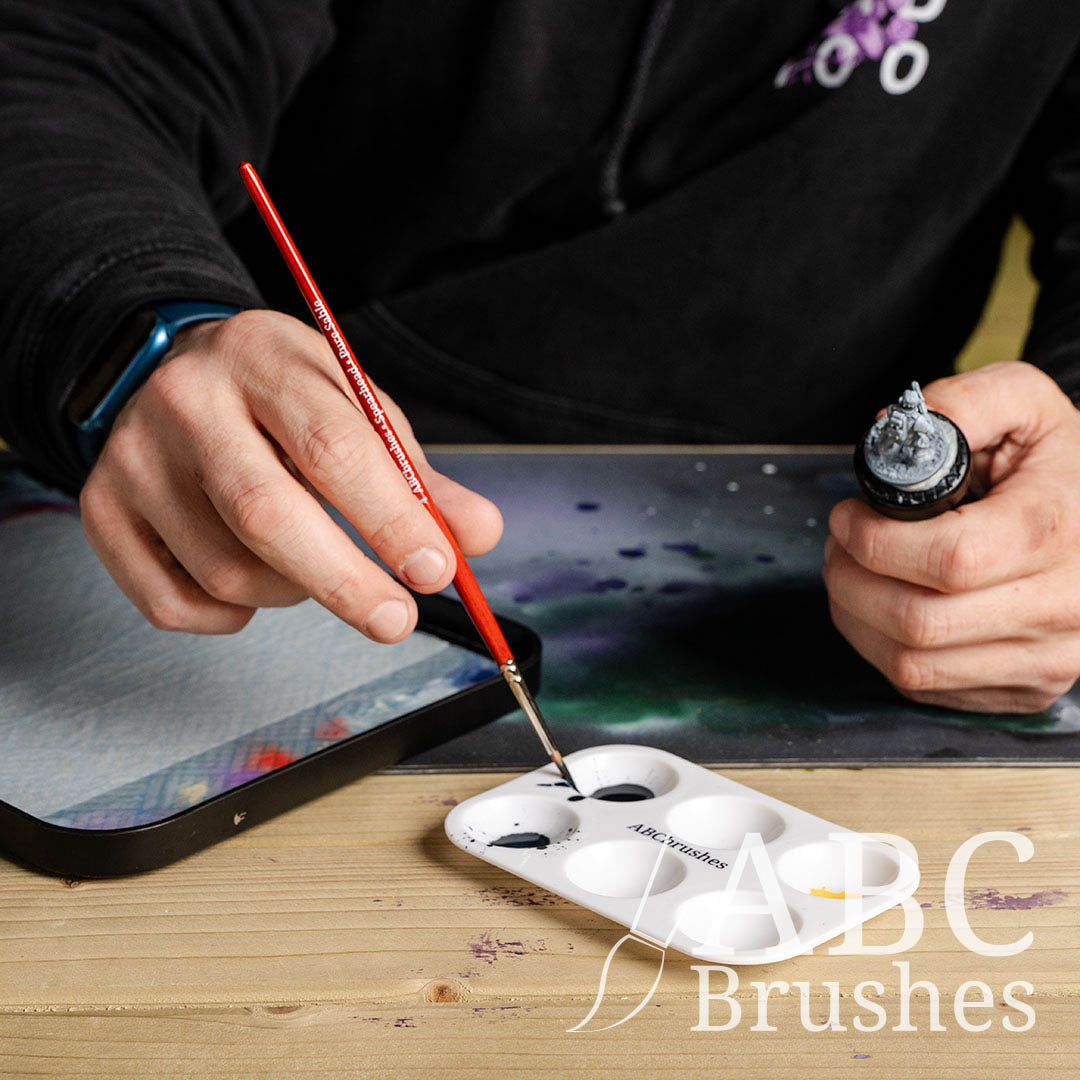 Person picking up paint with a ABC Brushes Spearhead paint brush with one hand and holding a mini in the other. There is a wet palette and cutting ma tin the background.