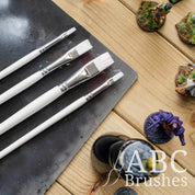 Close-up of the brush tips of the Pro Arte White Nylon flat paint brush set laid on top of a paint covered cutting mat and next to various mini figures. 