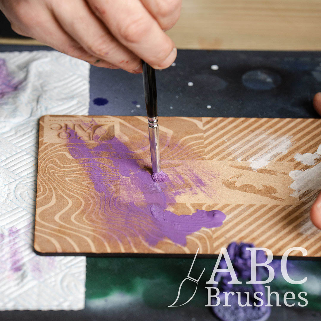 Removing purple paint from a Pro Arte dry brush using an ABC Brushes dry brush board, with a paper towel and purple miniature next to it. 