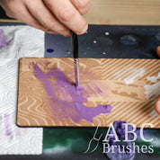 Removing purple paint from a Pro Arte dry brush using an ABC Brushes dry brush board, with a paper towel and purple miniature next to it. 