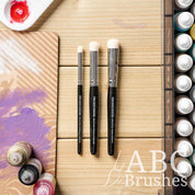The Army Painter Masterclass Drybrush Set on a wooden background, next to acrylic paint bottles, a drybrush board and a paint palette. 