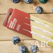 The Army Painters Most Wanted brush set packaging on a wooden background, next to various miniature figures. 