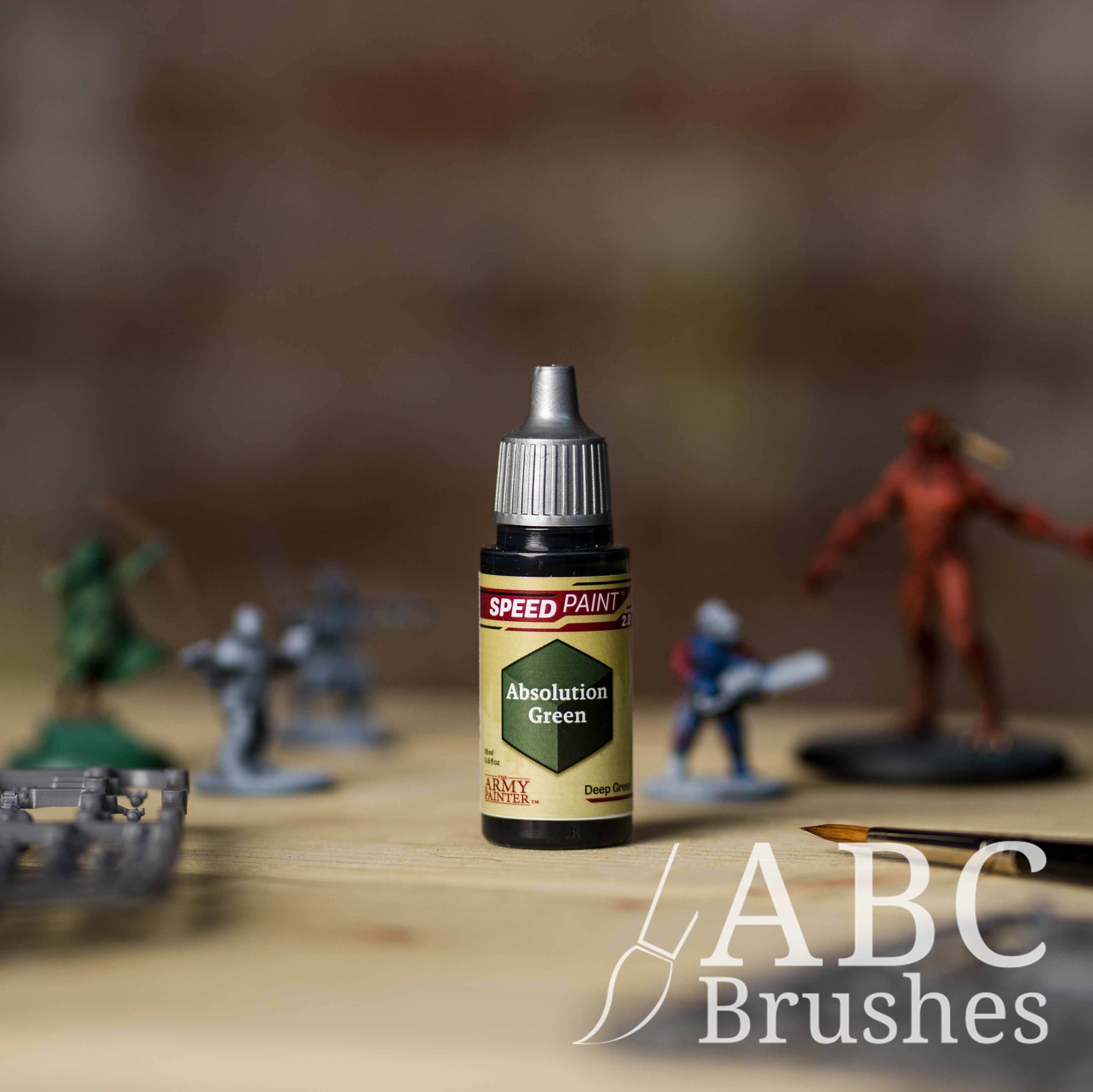 Bottle of the Army Painters Speedpaint in the colour Absolution Green, with wargaming models and an ABC paintbrush in the background, on a wooden surface.