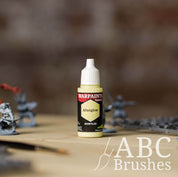 Bottle of the Army Painter Fanatics in the colour Afterglow with various wargaming models and an ABC paintbrush in the background, on a wooden surface.