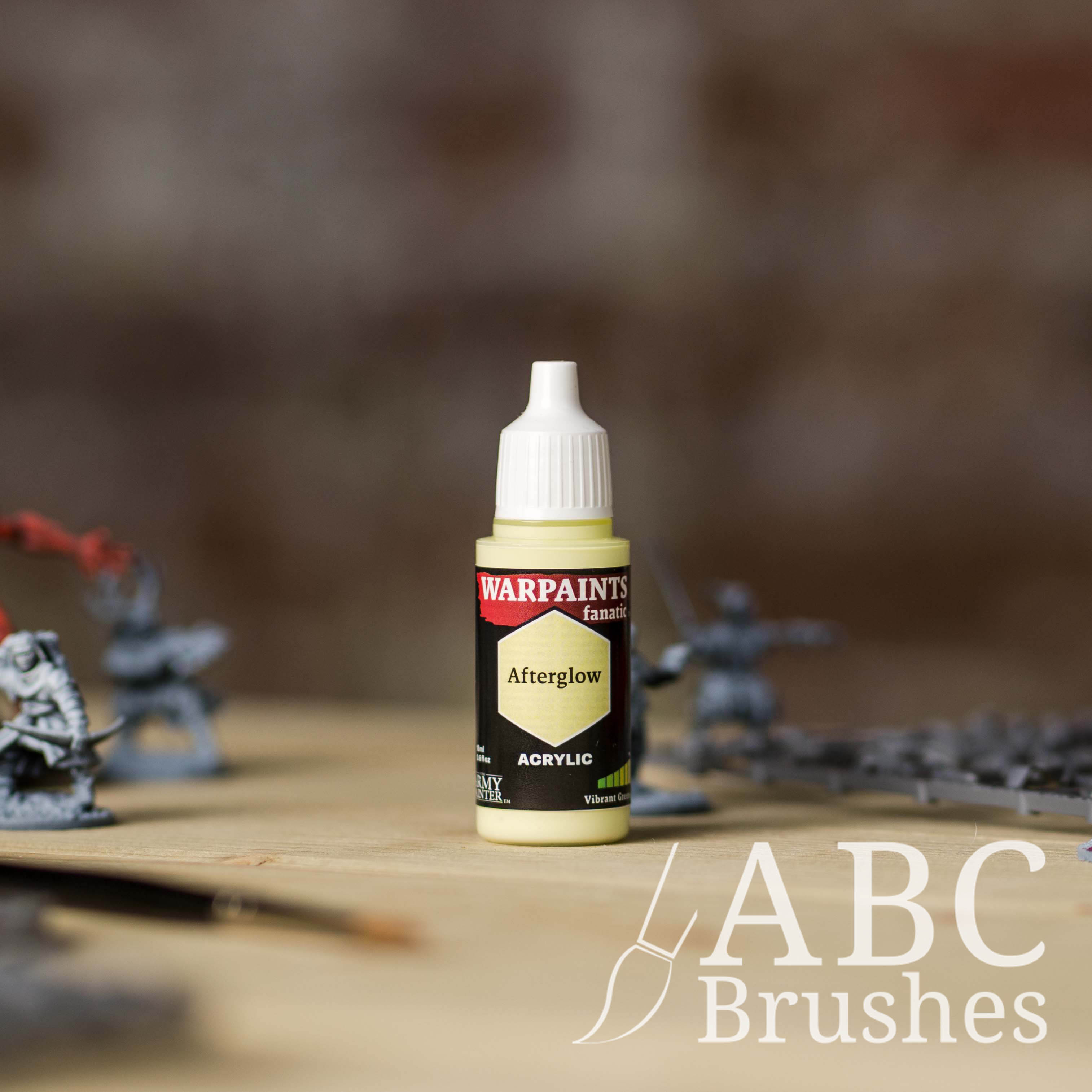 Bottle of the Army Painter Fanatics in the colour Afterglow with various wargaming models and an ABC paintbrush in the background, on a wooden surface.