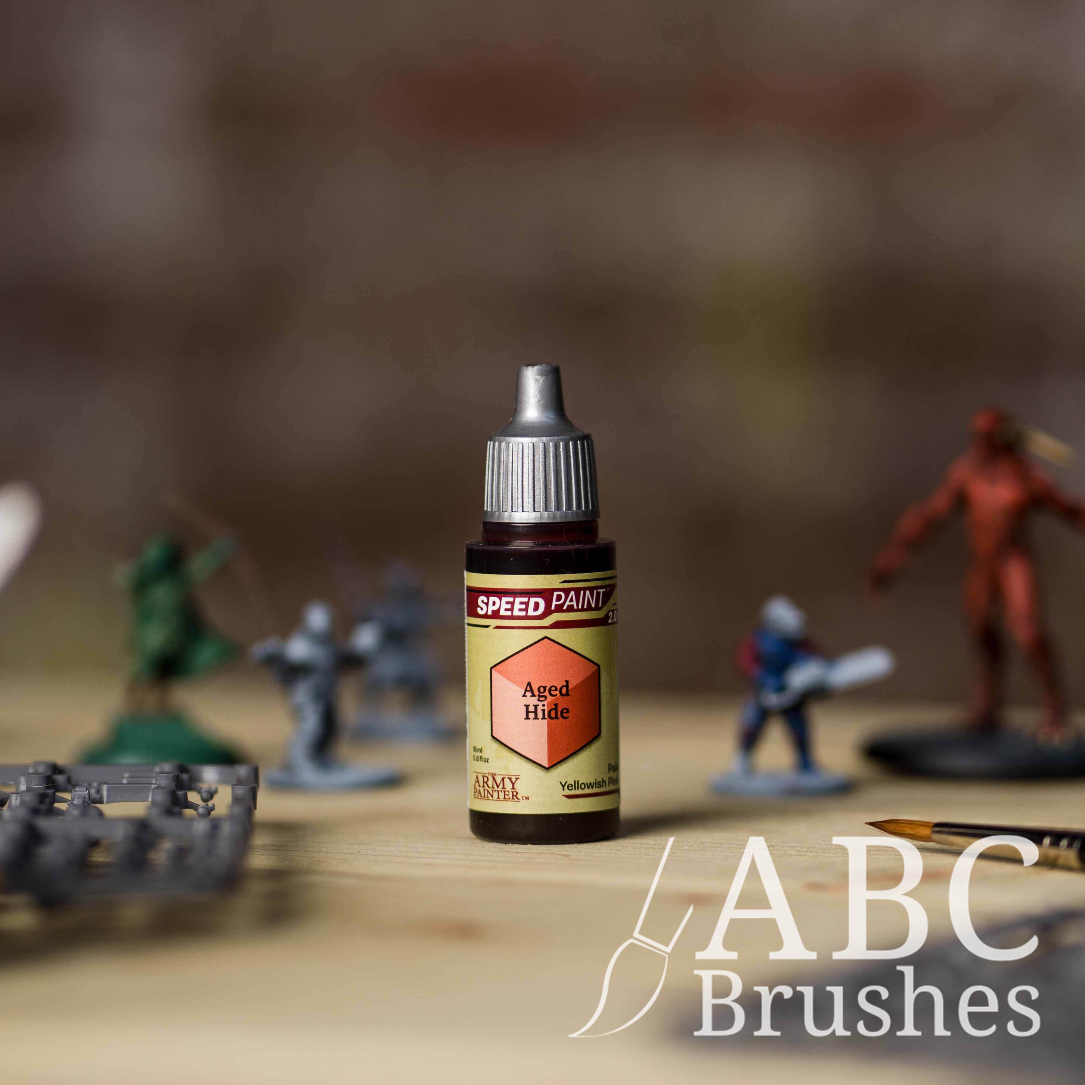 Bottle of the Army Painters Speedpaint in the colour Aged Hide, with wargaming models and an ABC paintbrush in the background, on a wooden surface.