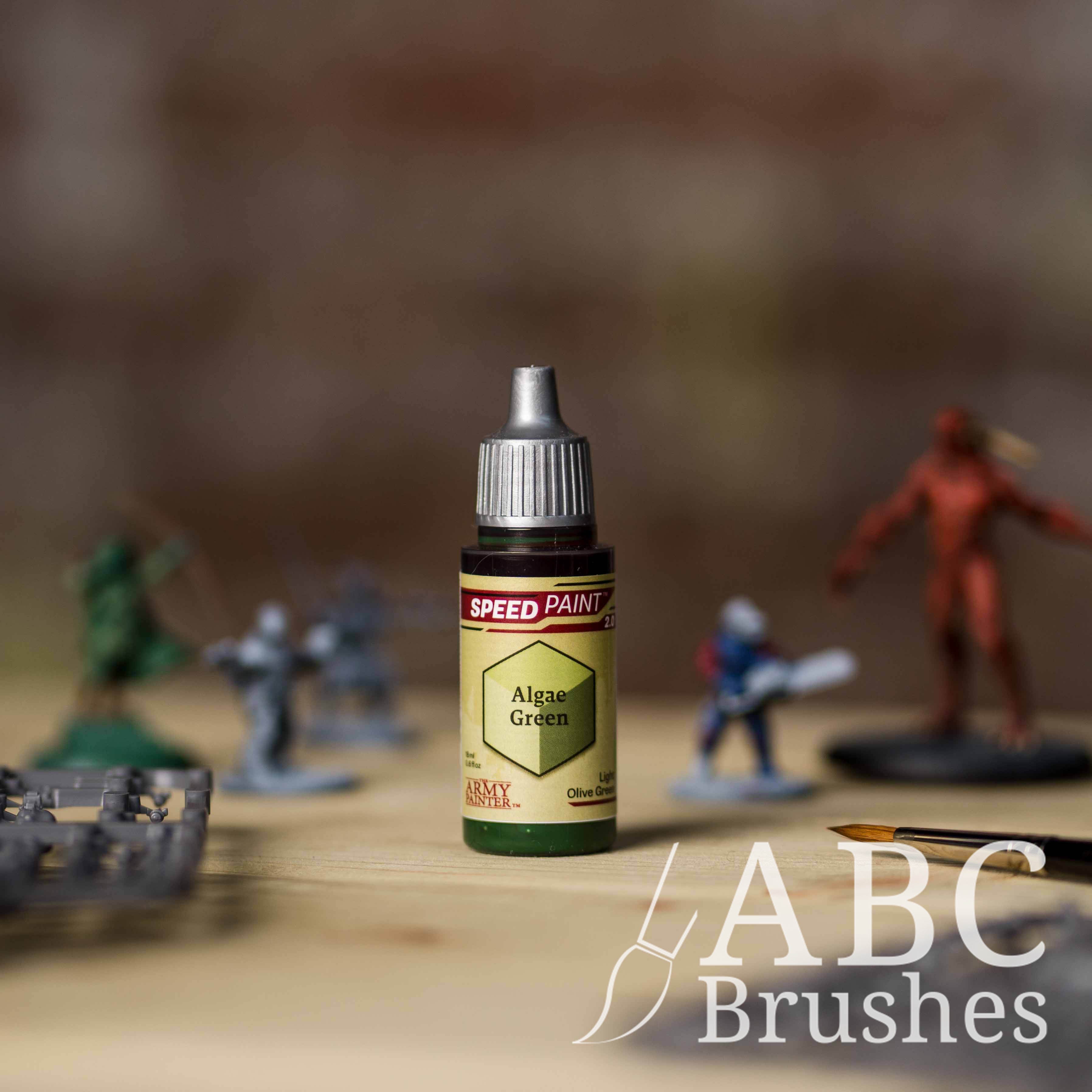 Bottle of the Army Painters Speedpaint in the colour Algae Green, with wargaming models and an ABC paintbrush in the background, on a wooden surface.