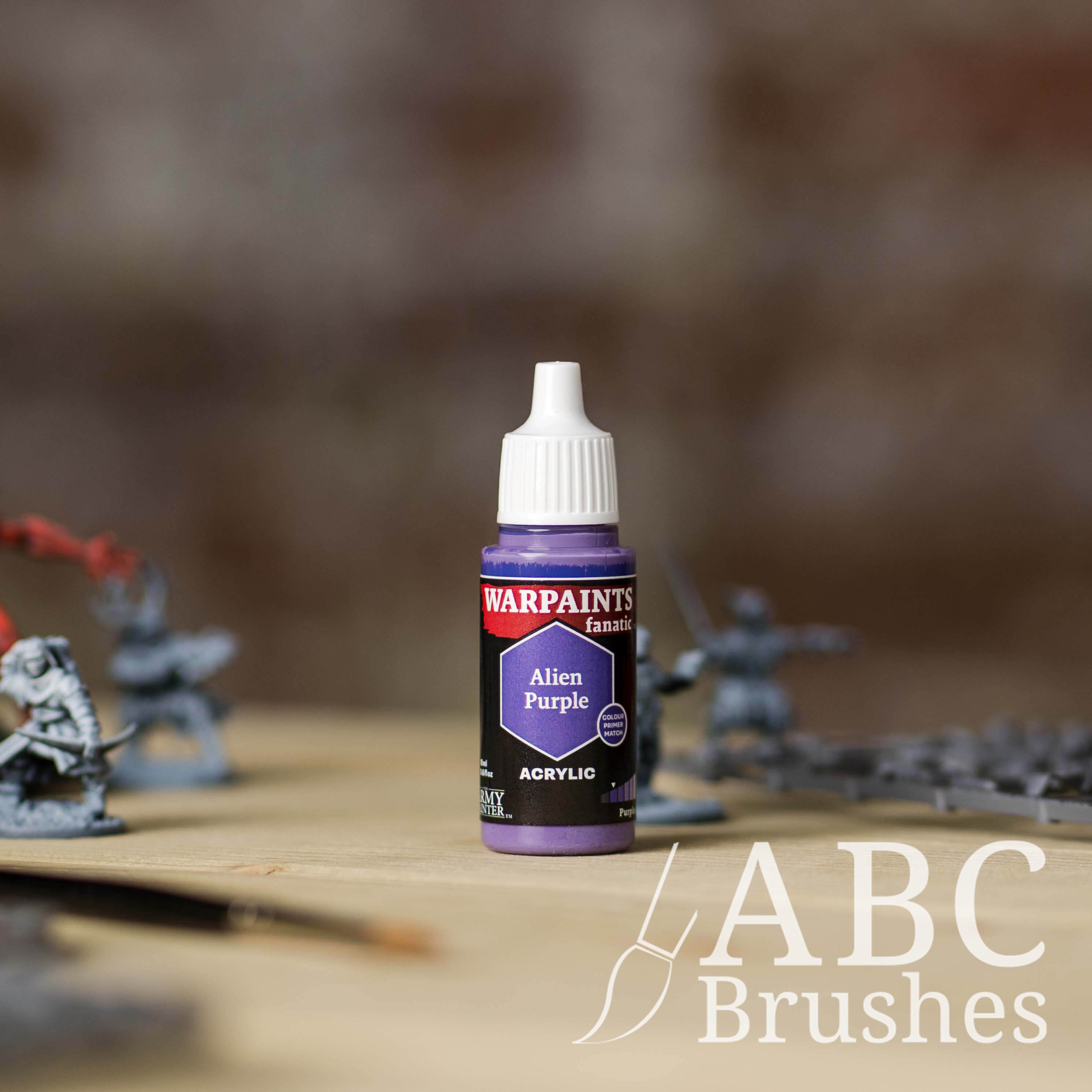 Bottle of the Army Painter Fanatics in the colour Alien Purple with various wargaming models and an ABC paintbrush in the background, on a wooden surface.