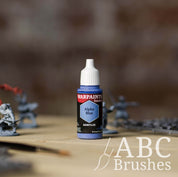 Bottle of the Army Painter Fanatics in the colour Alpha Blue with various wargaming models and an ABC paintbrush in the background, on a wooden surface.