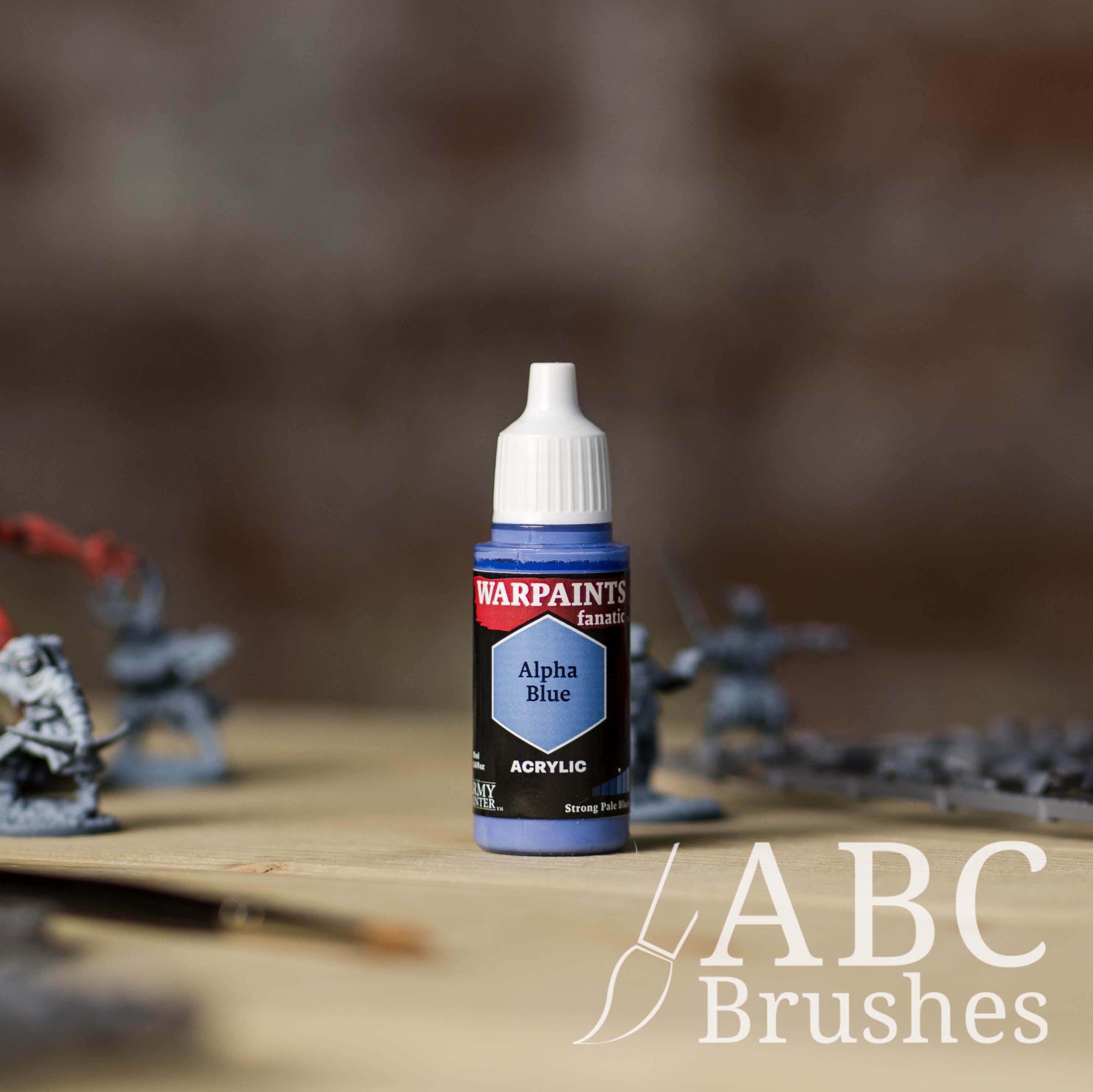 Bottle of the Army Painter Fanatics in the colour Alpha Blue with various wargaming models and an ABC paintbrush in the background, on a wooden surface.