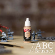 Bottle of the Army Painter Fanatics in the colour Amber Skin with various wargaming models and an ABC paintbrush in the background, on a wooden surface.