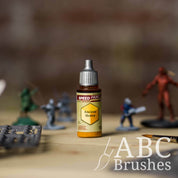 Bottle of the Army Painters Speedpaint in the colour Ancient Honey, with wargaming models and an ABC paintbrush in the background, on a wooden surface.