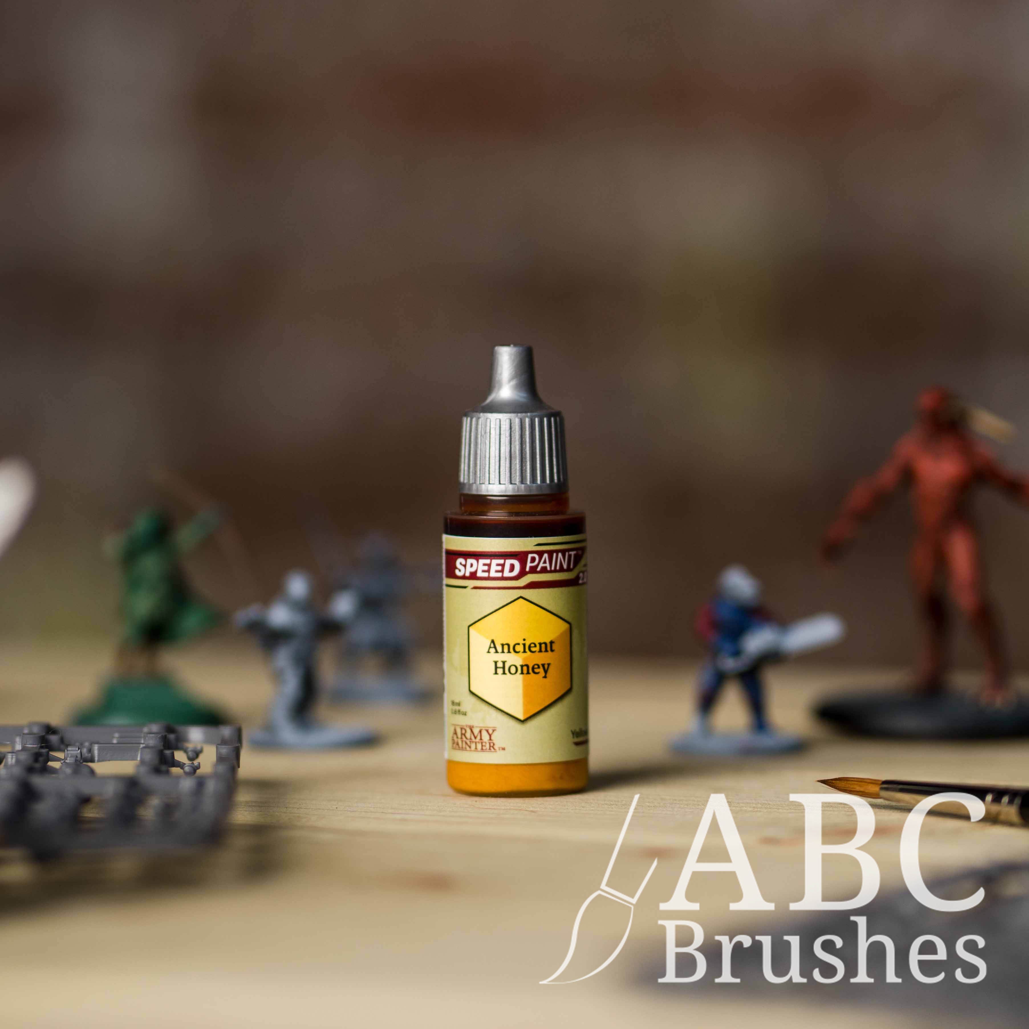 Bottle of the Army Painters Speedpaint in the colour Ancient Honey, with wargaming models and an ABC paintbrush in the background, on a wooden surface.