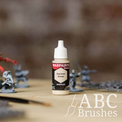 Bottle of the Army Painter Fanatics in the colour Ancient Stone with various wargaming models and an ABC paintbrush in the background, on a wooden surface.