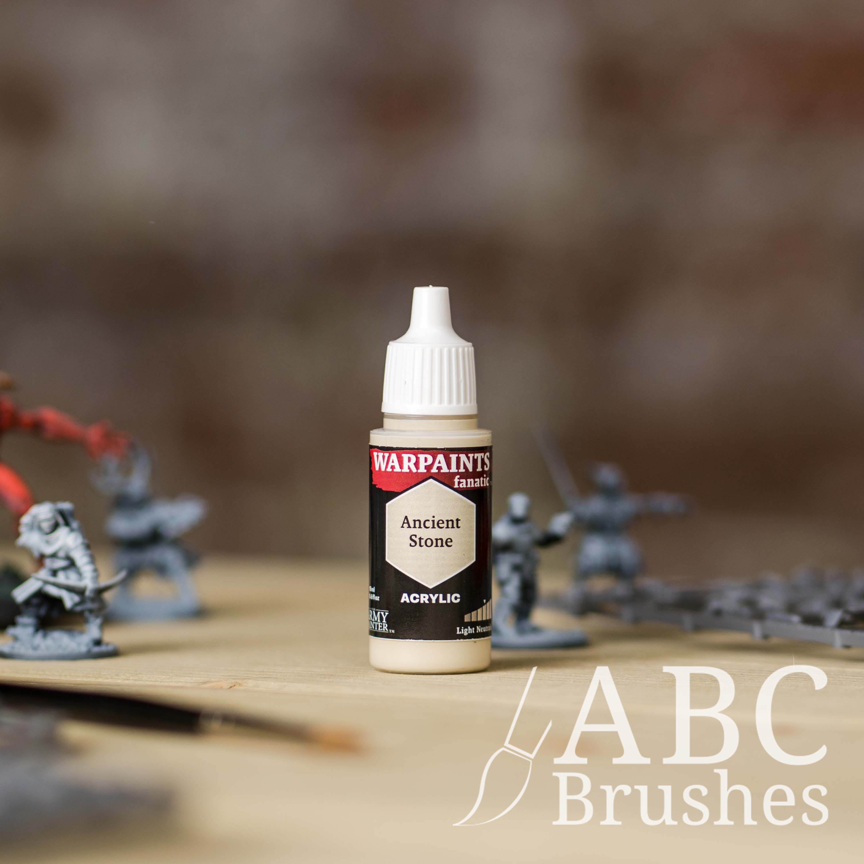 Bottle of the Army Painter Fanatics in the colour Ancient Stone with various wargaming models and an ABC paintbrush in the background, on a wooden surface.