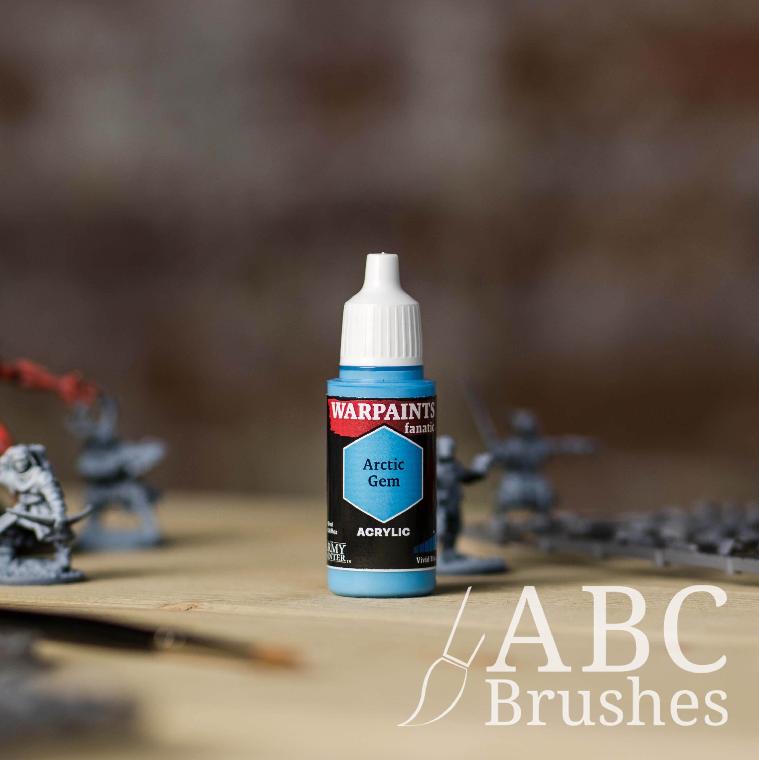 Bottle of the Army Painter Fanatics in the colour Arctic Gem with various wargaming models and an ABC paintbrush in the background, on a wooden surface.