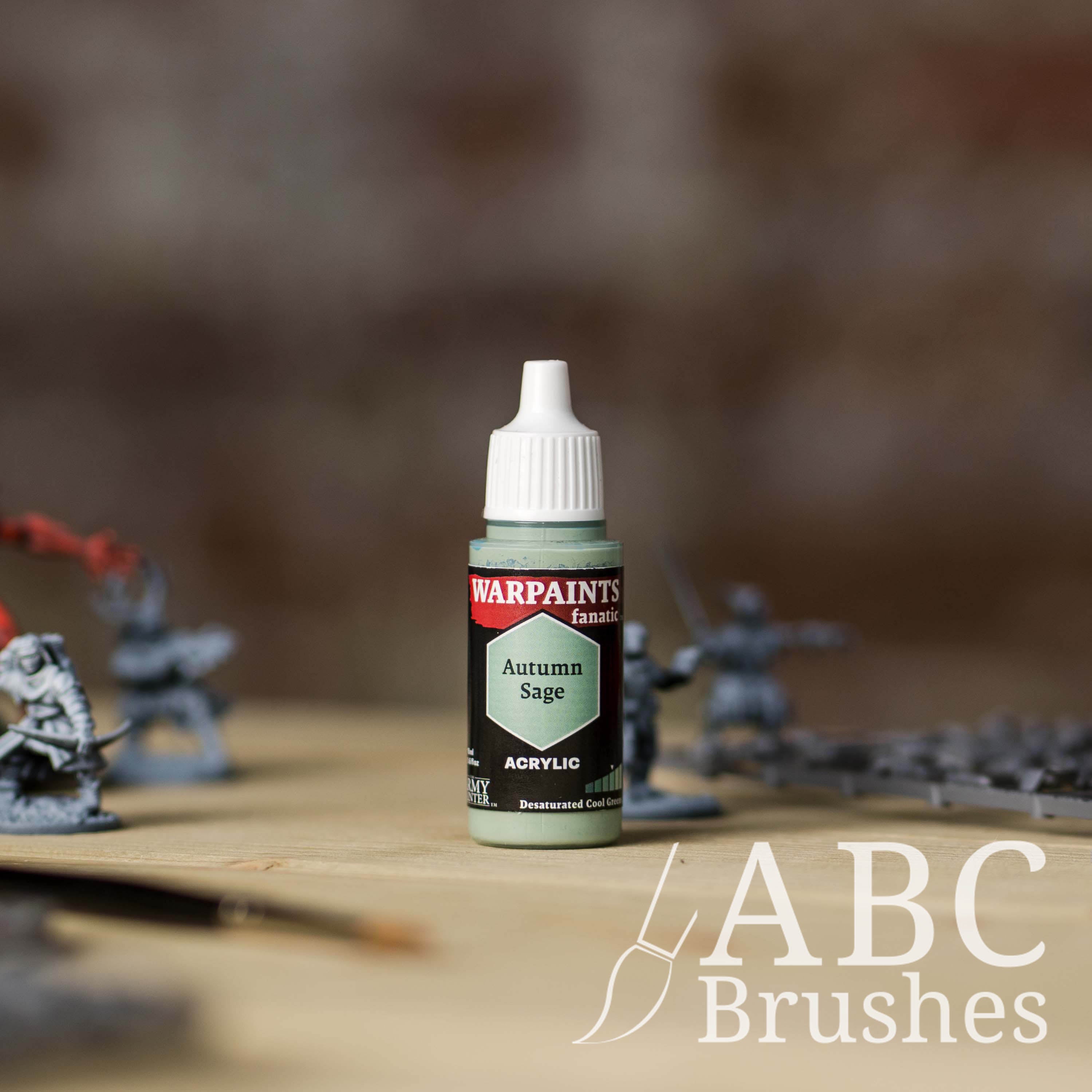 Bottle of the Army Painter Fanatics in the colour Autumn Sage with various wargaming models and an ABC paintbrush in the background, on a wooden surface.