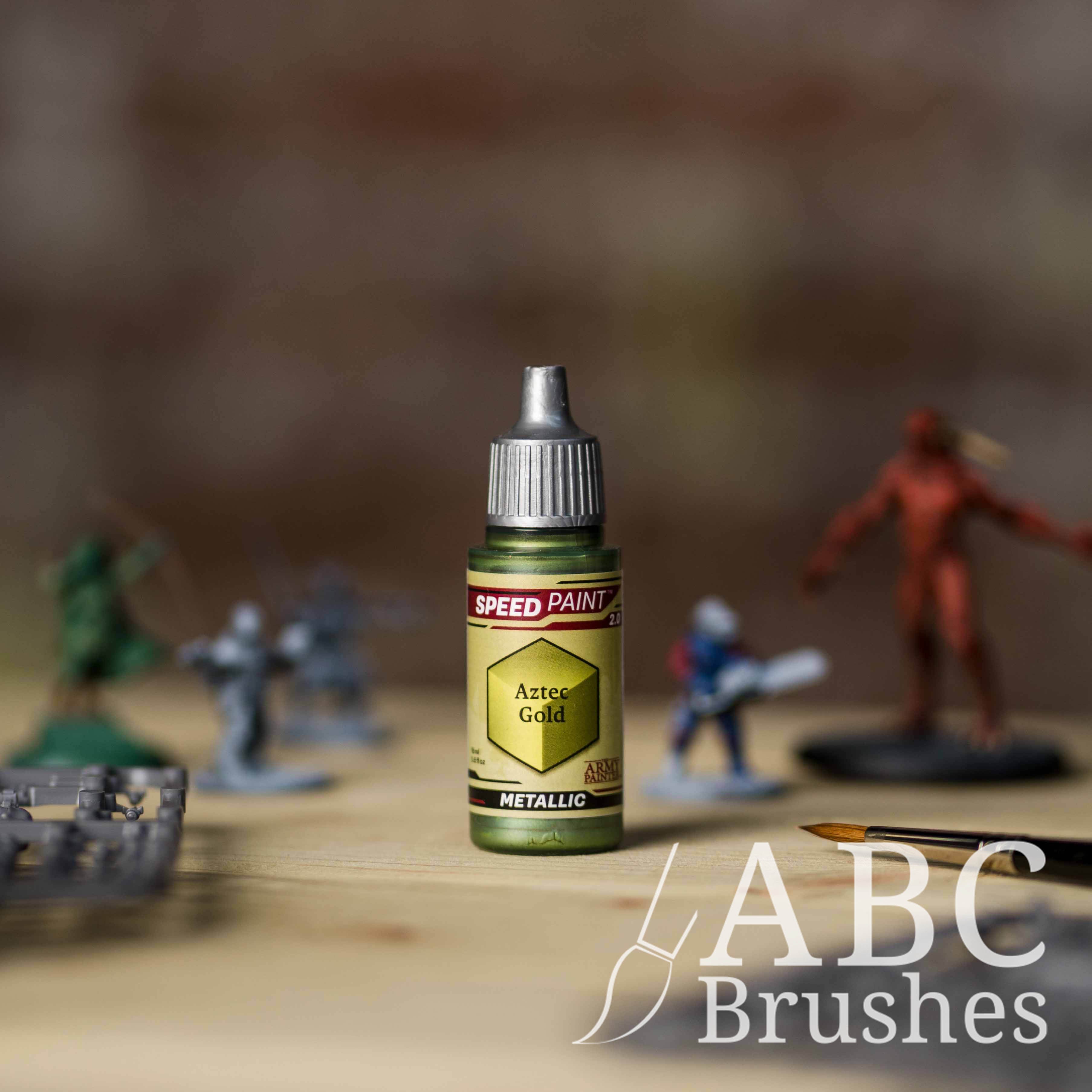 Bottle of the Army Painters Metallic Speedpaint in the colour Aztec Gold, with wargaming models and an ABC paintbrush in the background, on a wooden surface.