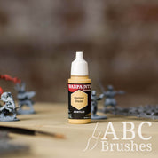 Bottle of the Army Painter Fanatics in the colour Barren Dune with various wargaming models and an ABC paintbrush in the background, on a wooden surface.