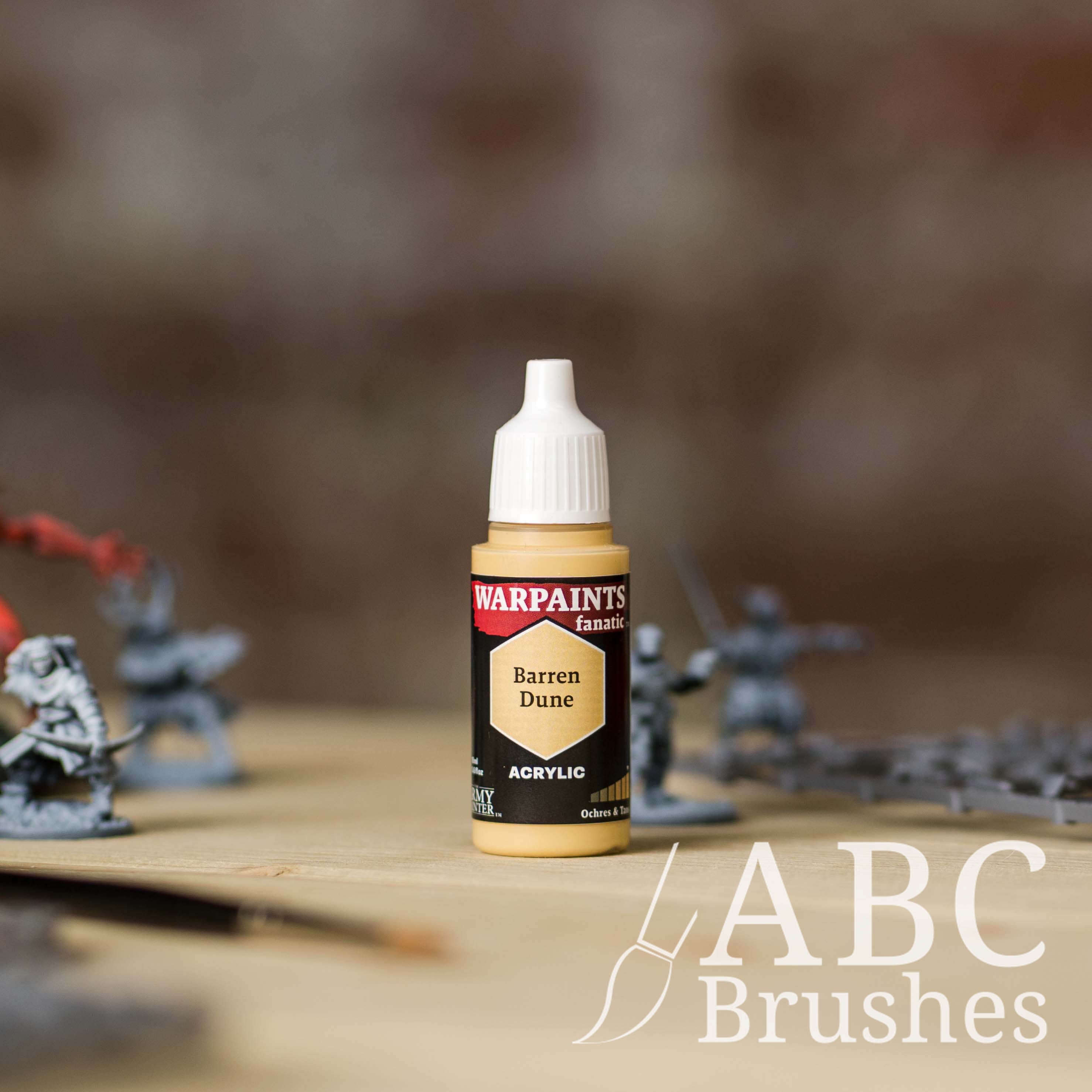 Bottle of the Army Painter Fanatics in the colour Barren Dune with various wargaming models and an ABC paintbrush in the background, on a wooden surface.