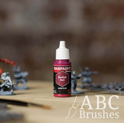 Bottle of the Army Painter Fanatics in the colour Basilisk Red with various wargaming models and an ABC paintbrush in the background, on a wooden surface.