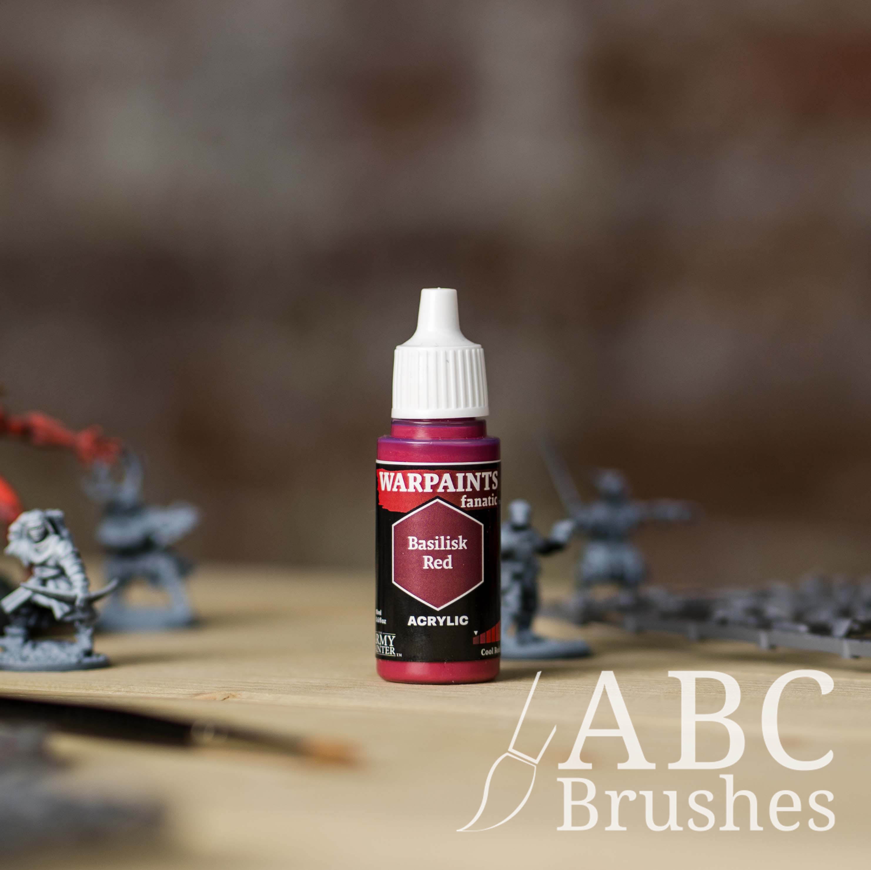Bottle of the Army Painter Fanatics in the colour Basilisk Red with various wargaming models and an ABC paintbrush in the background, on a wooden surface.