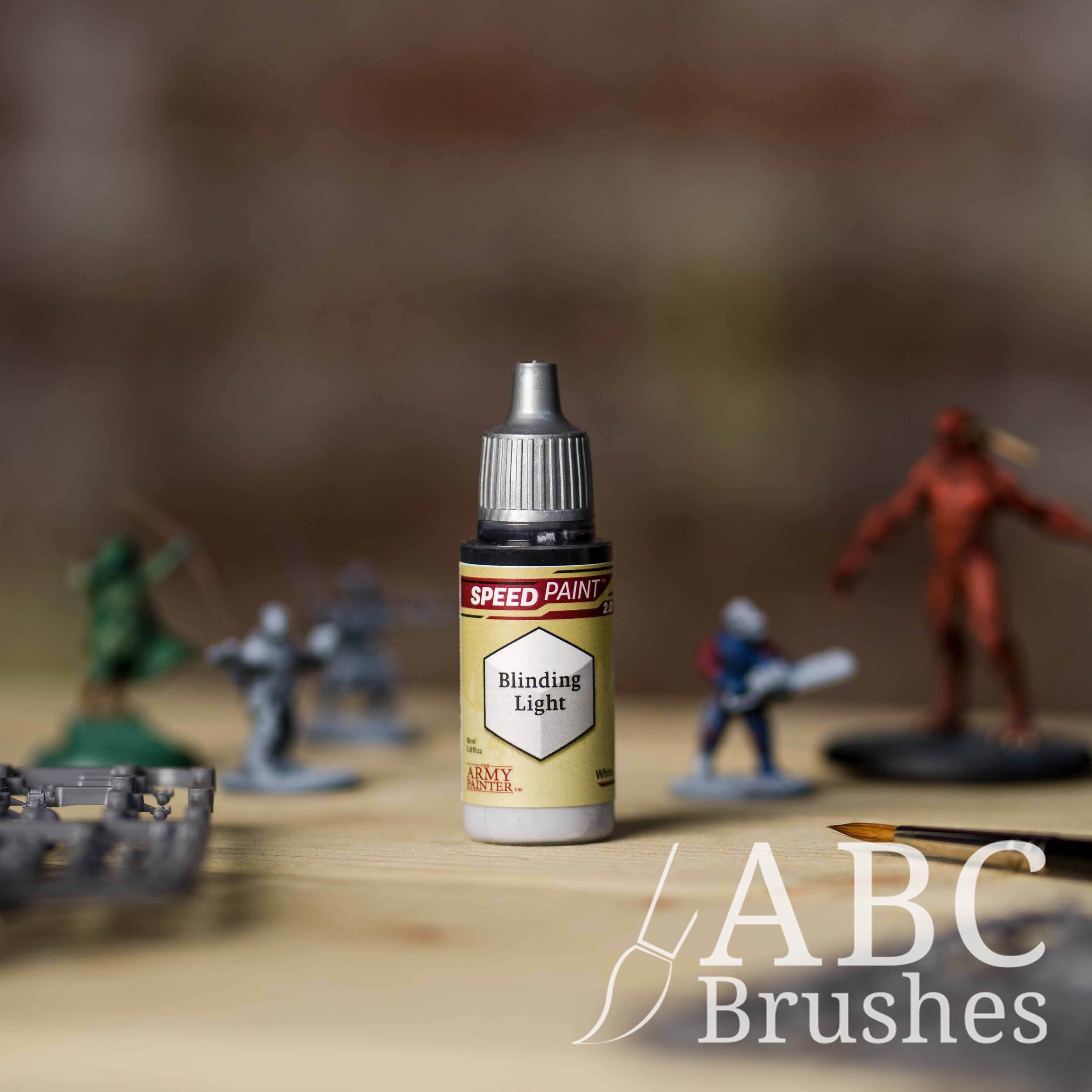 Bottle of the Army Painters Speedpaint in the colour Blinding Light, with wargaming models and an ABC paintbrush in the background, on a wooden surface.