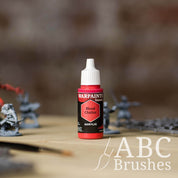 Bottle of the Army Painter Fanatics in the colour Blood Chalice with various wargaming models and an ABC paintbrush in the background, on a wooden surface.