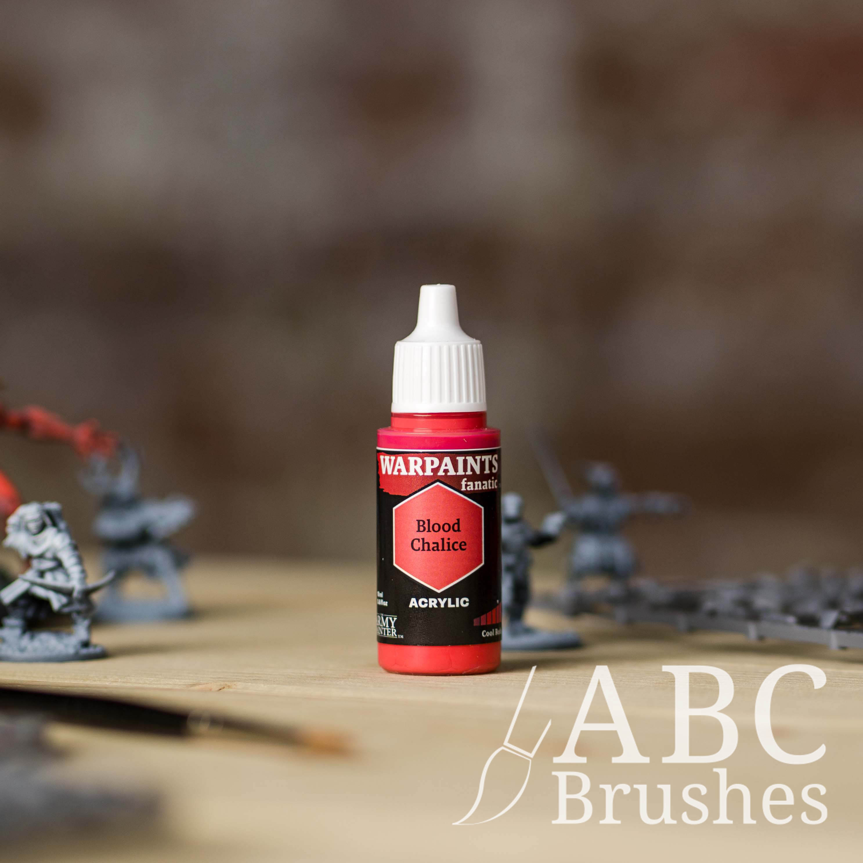 Bottle of the Army Painter Fanatics in the colour Blood Chalice with various wargaming models and an ABC paintbrush in the background, on a wooden surface.