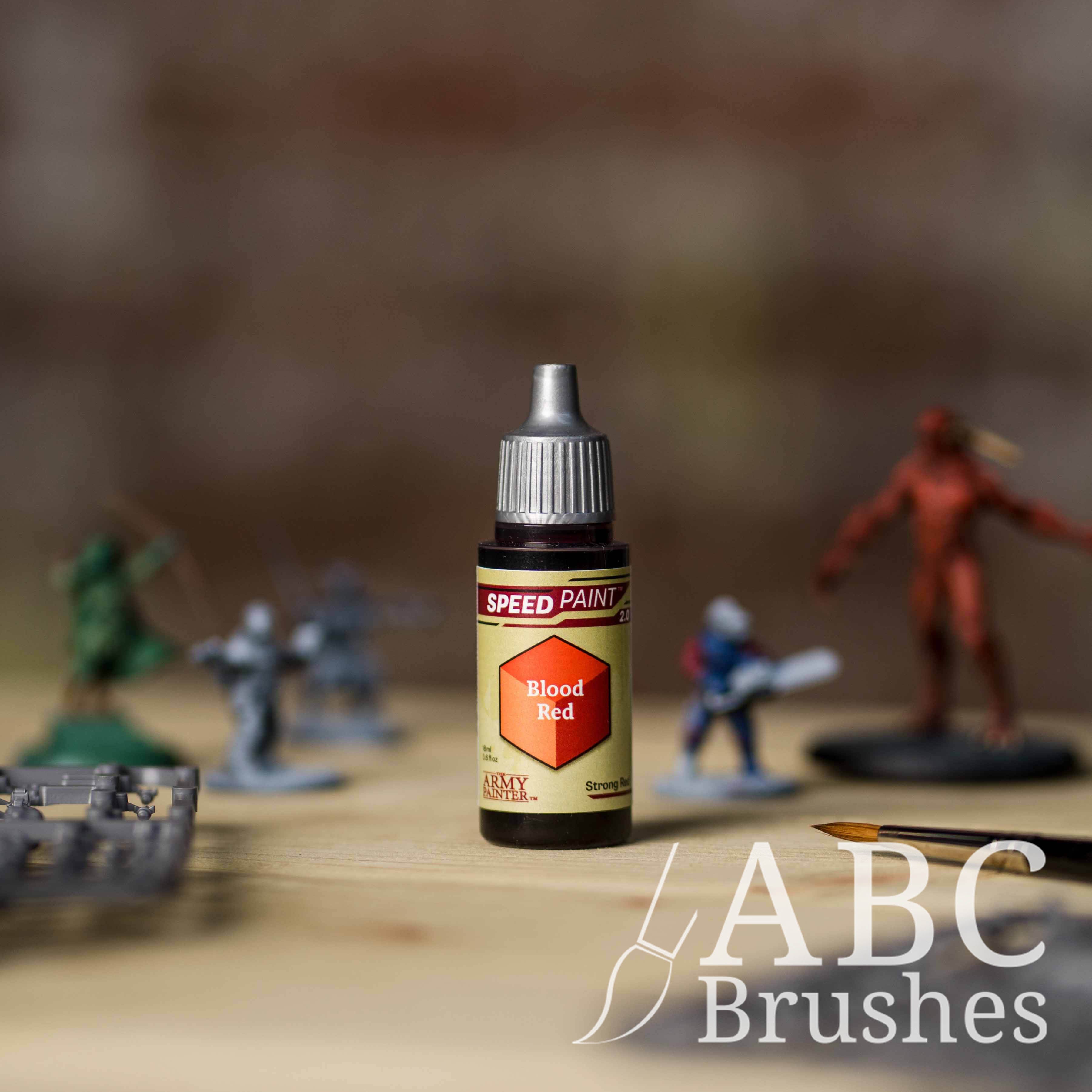 Bottle of the Army Painters Speedpaint in the colour Blood Red, with wargaming models and an ABC paintbrush in the background, on a wooden surface.