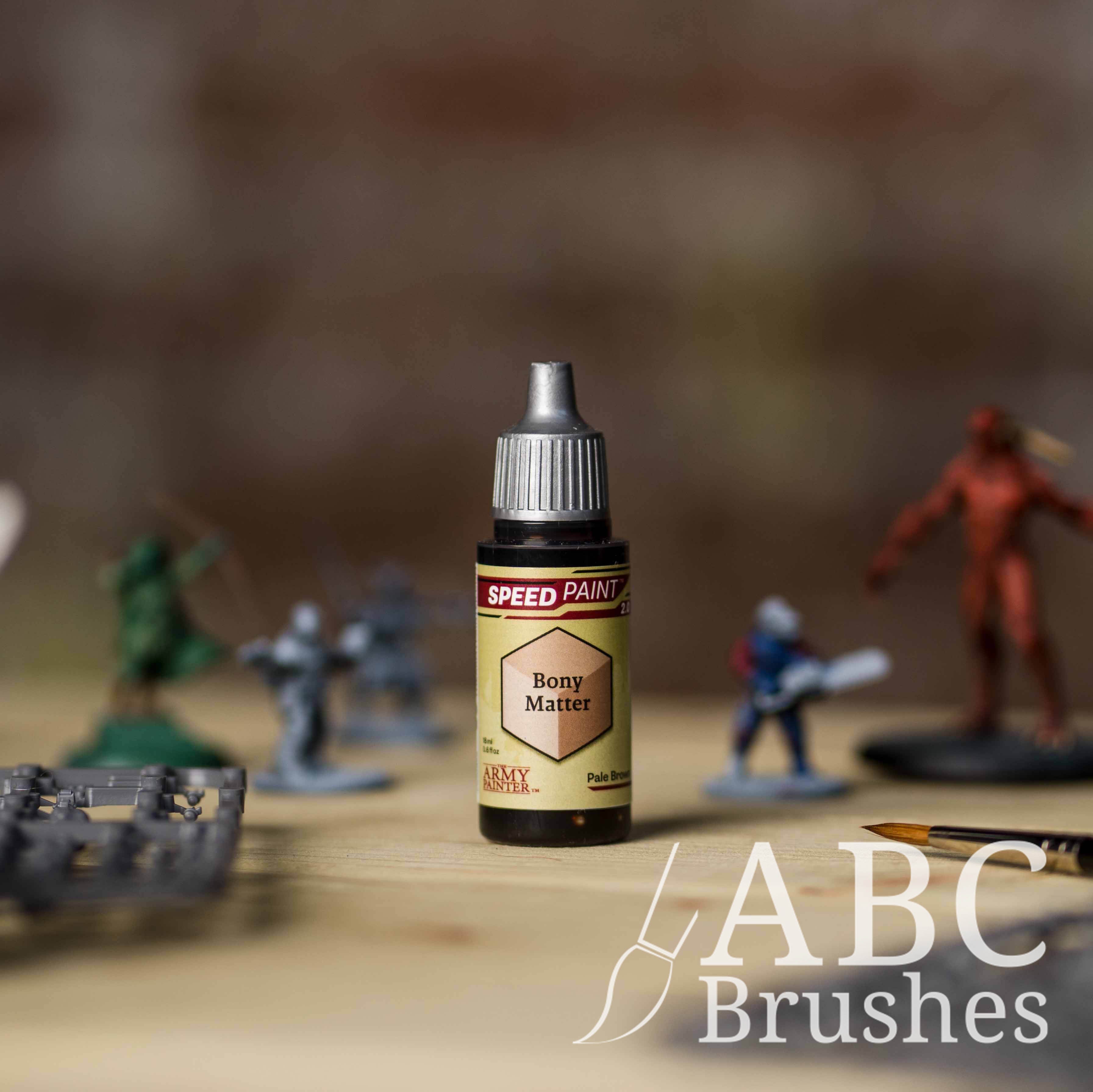 Bottle of the Army Painters Speedpaint in the colour Bony Matter, with wargaming models and an ABC paintbrush in the background, on a wooden surface.