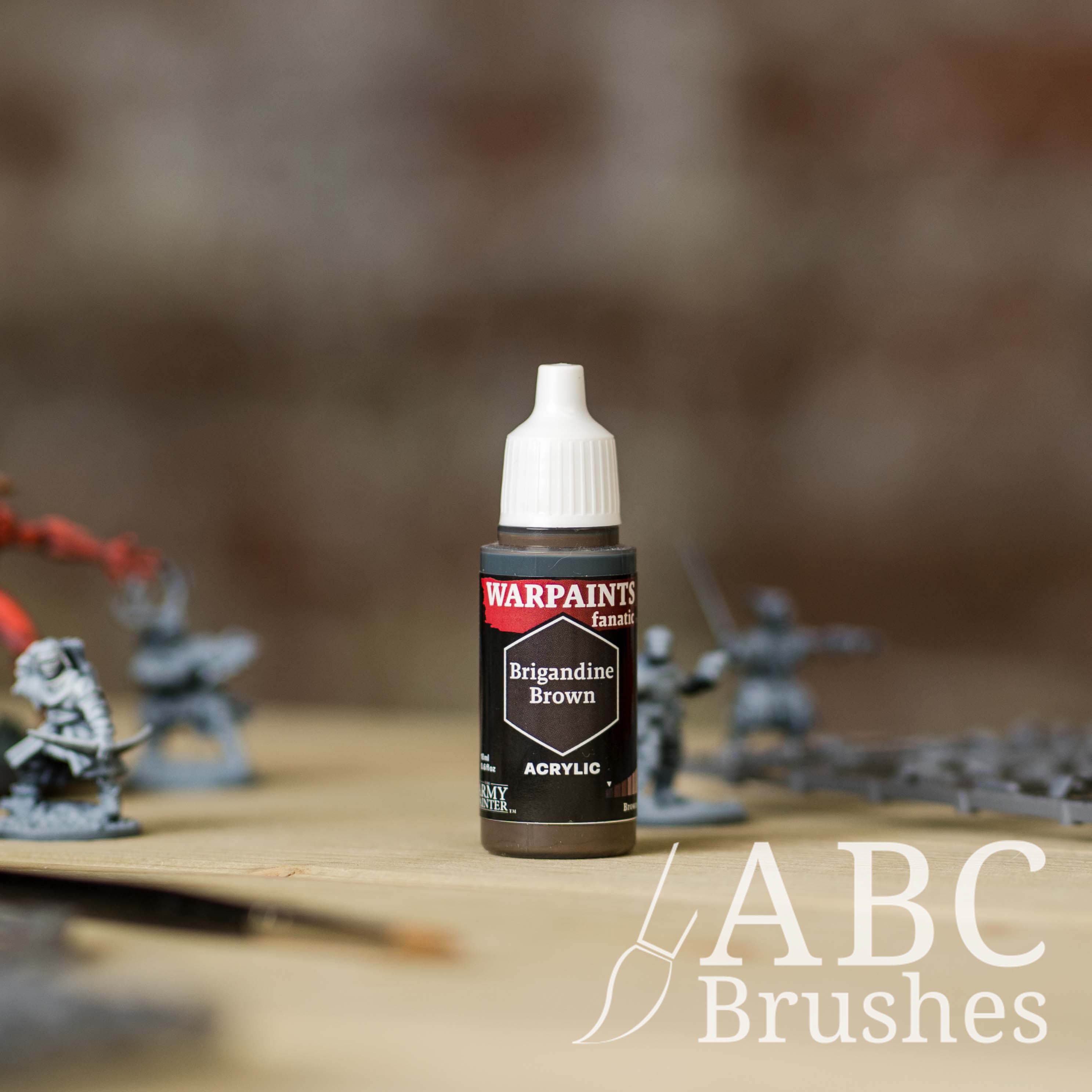 Bottle of the Army Painter Fanatics in the colour Brigandine Brown with various wargaming models and an ABC paintbrush in the background, on a wooden surface.