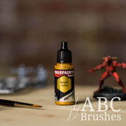 Bottle of the Army Painter Fanatics Metallic paint in the colour Bright Gold with various wargaming models and an ABC paintbrush in the background, on a wooden surface.