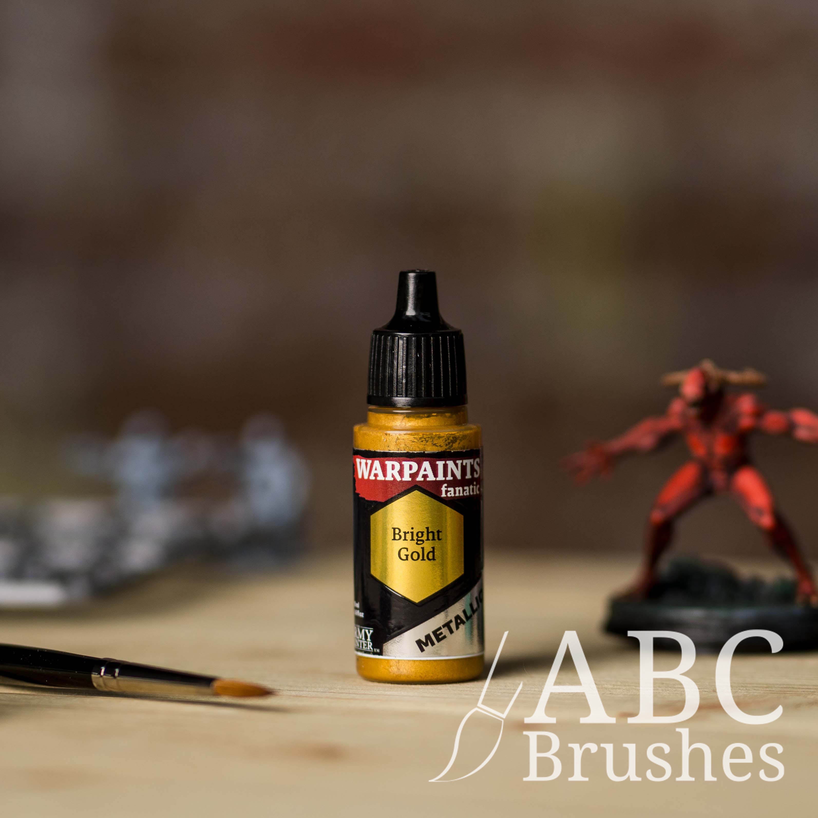 Bottle of the Army Painter Fanatics Metallic paint in the colour Bright Gold with various wargaming models and an ABC paintbrush in the background, on a wooden surface.
