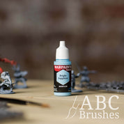 Bottle of the Army Painter Fanatics in the colour Bright Sapphire with various wargaming models and an ABC paintbrush in the background, on a wooden surface.