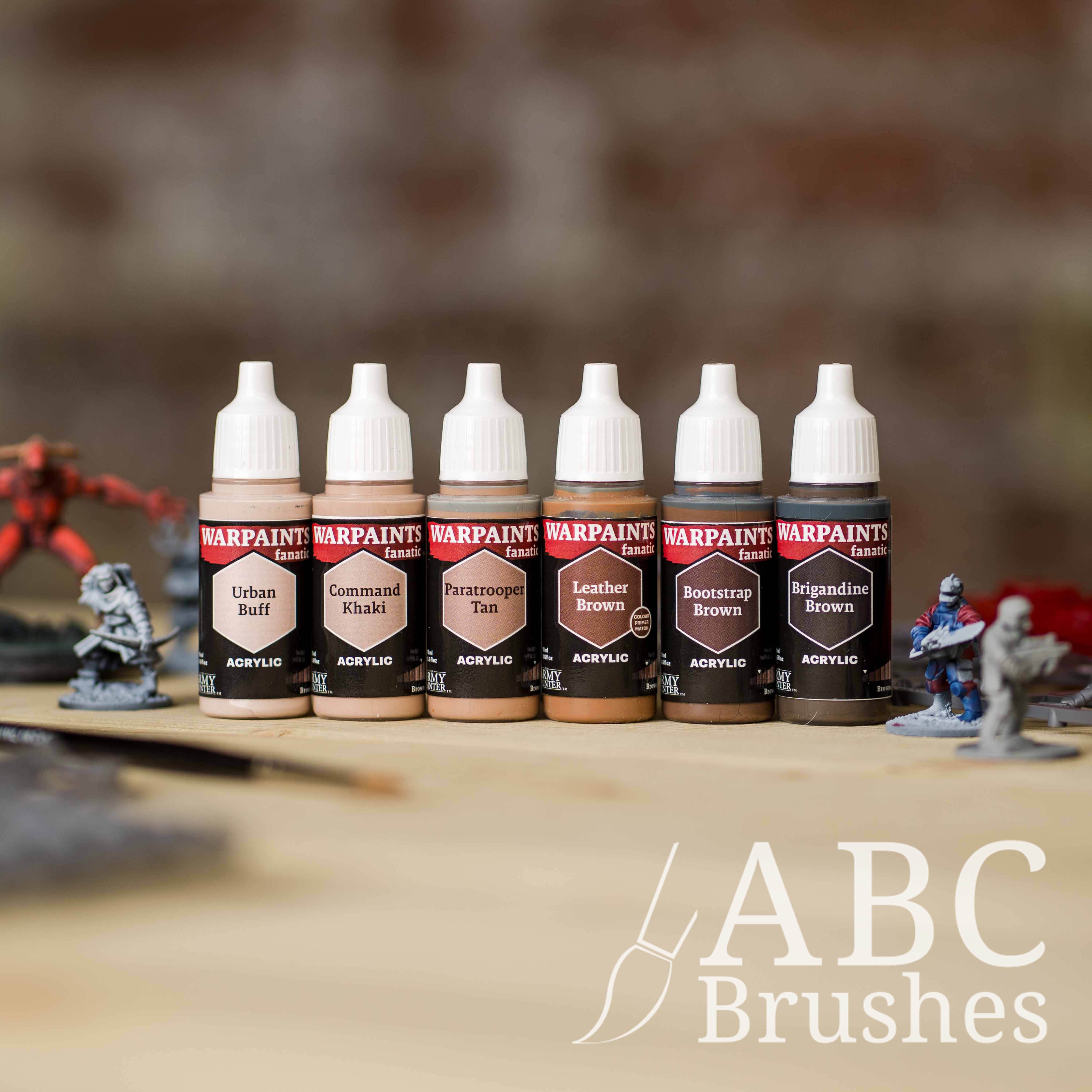 Army Painter Fanatics Browns Triad on a wooden surface. There are wargaming models and an ABC paint brush next to them.