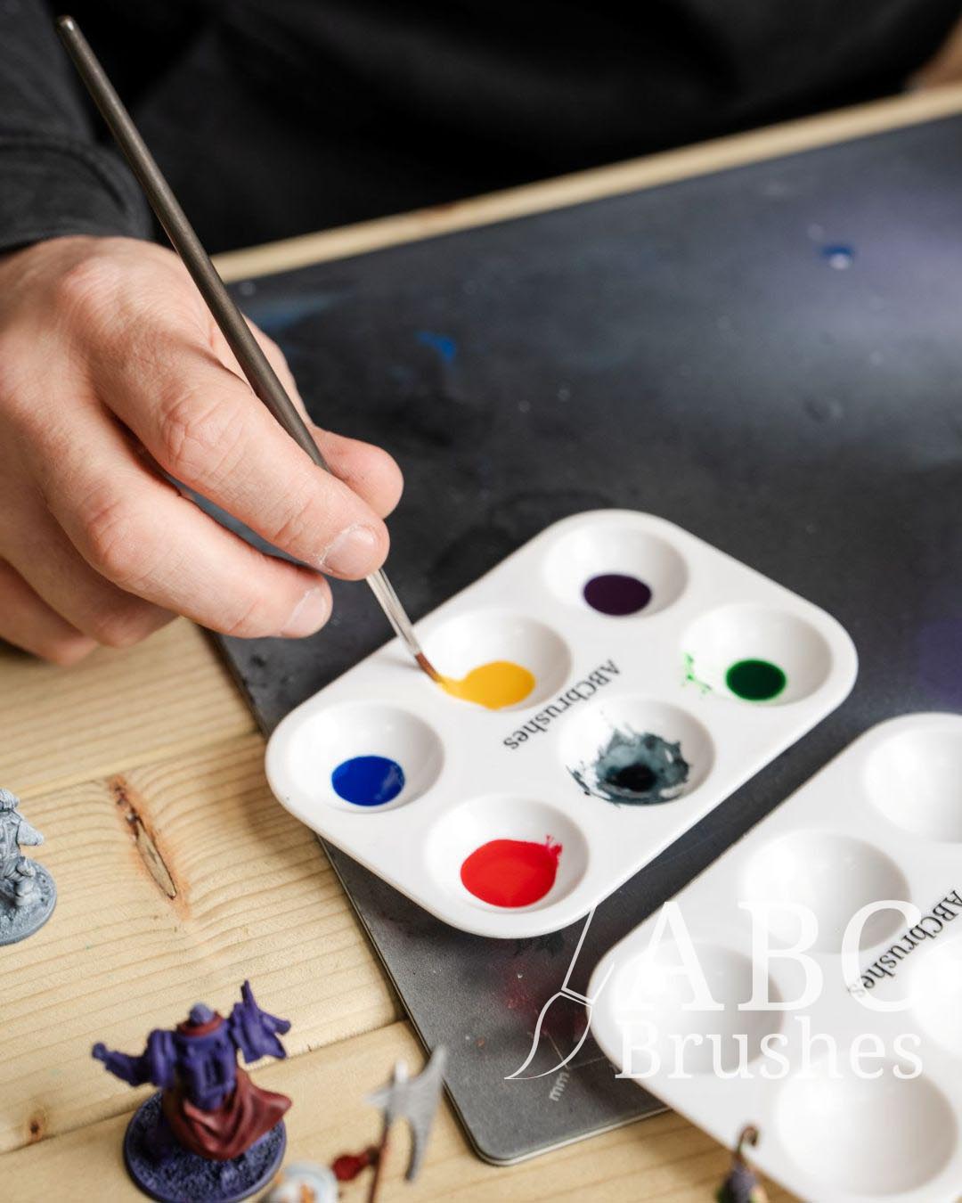Picking up yellow paint with a round paintbrush, from an abc paint palette. There are minis next to it. 