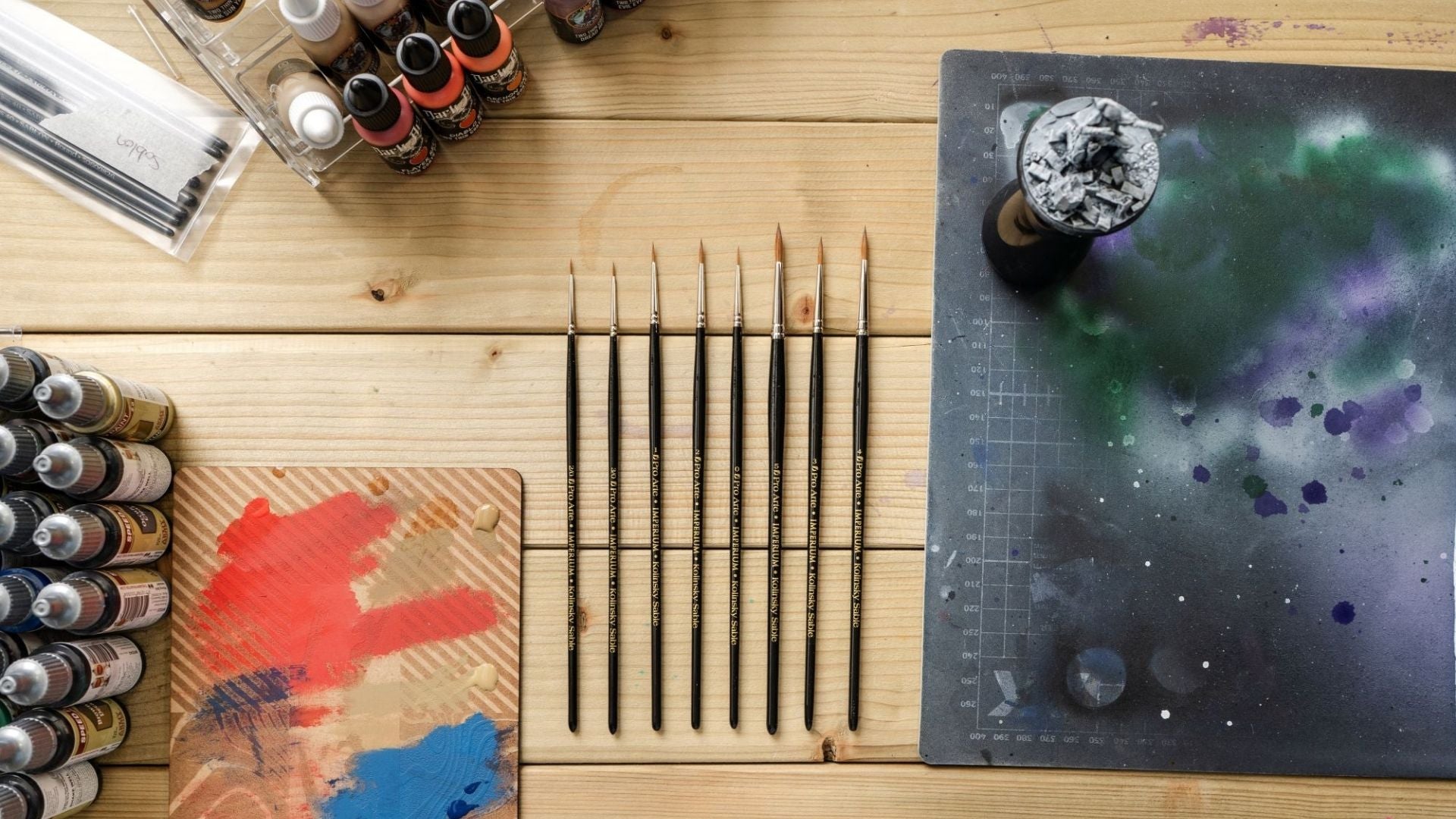 An overhead flat-lay of an miniature painter's paint setup on a wooden table, featuring multiple paintbrushes, a dry brush board and bottles of acrylic paint.