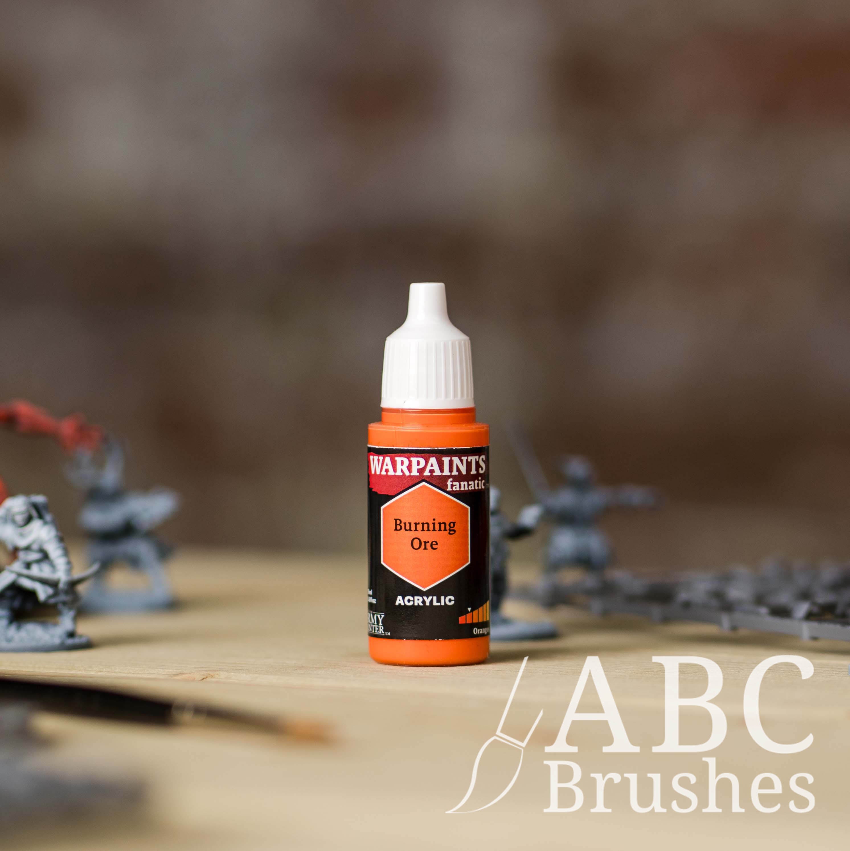 Bottle of the Army Painter Fanatics in the colour Burning Ore with various wargaming models and an ABC paintbrush in the background, on a wooden surface.