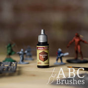 Bottle of the Army Painters Speedpaint in the colour Burnished Red, with wargaming models and an ABC paintbrush in the background, on a wooden surface.