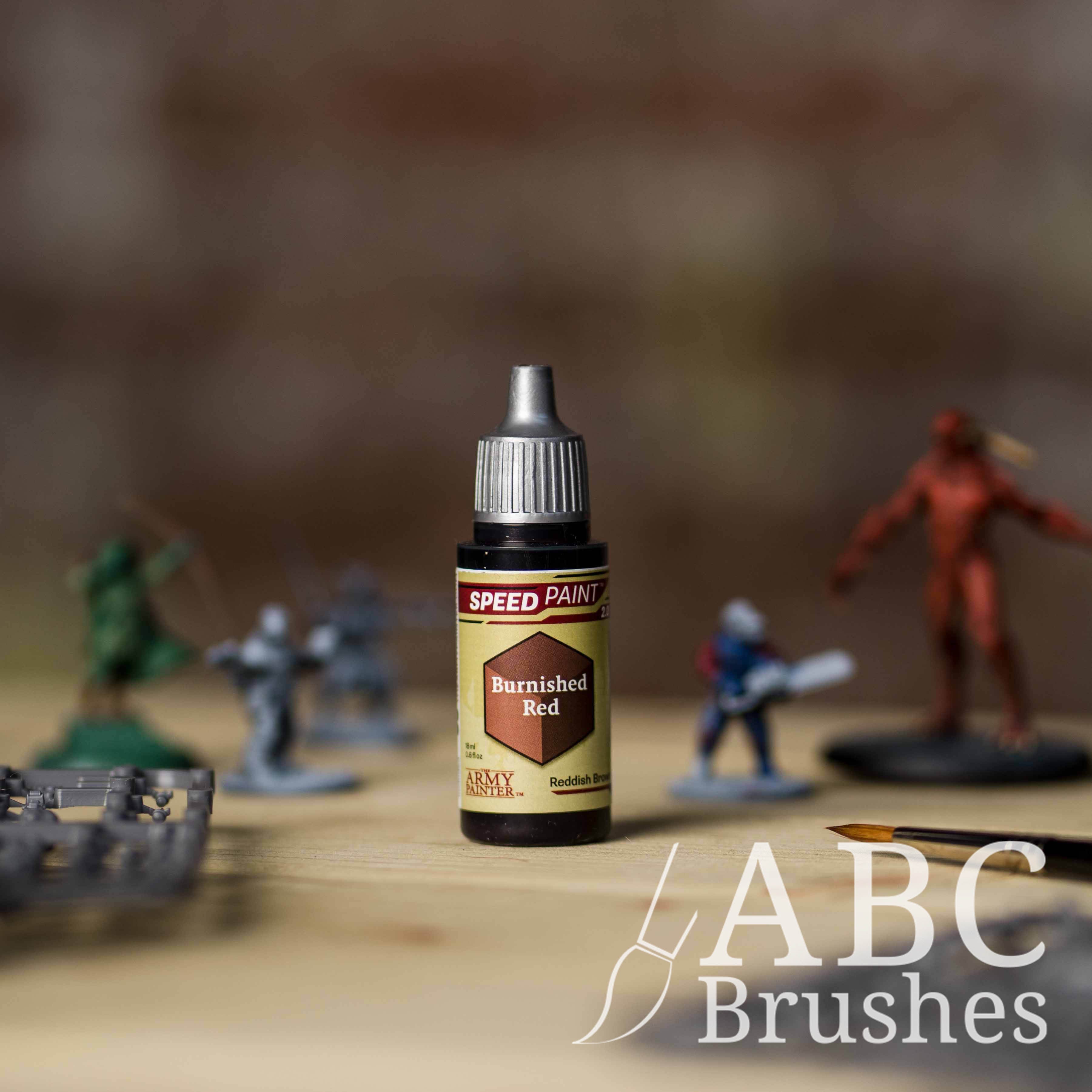 Bottle of the Army Painters Speedpaint in the colour Burnished Red, with wargaming models and an ABC paintbrush in the background, on a wooden surface.