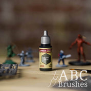 Bottle of the Army Painters Speedpaint in the colour Burnt Moss, with wargaming models and an ABC paintbrush in the background, on a wooden surface.