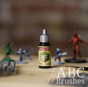 Bottle of the Army Painters Speedpaint in the colour Camo Cloak, with wargaming models and an ABC paintbrush in the background, on a wooden surface.