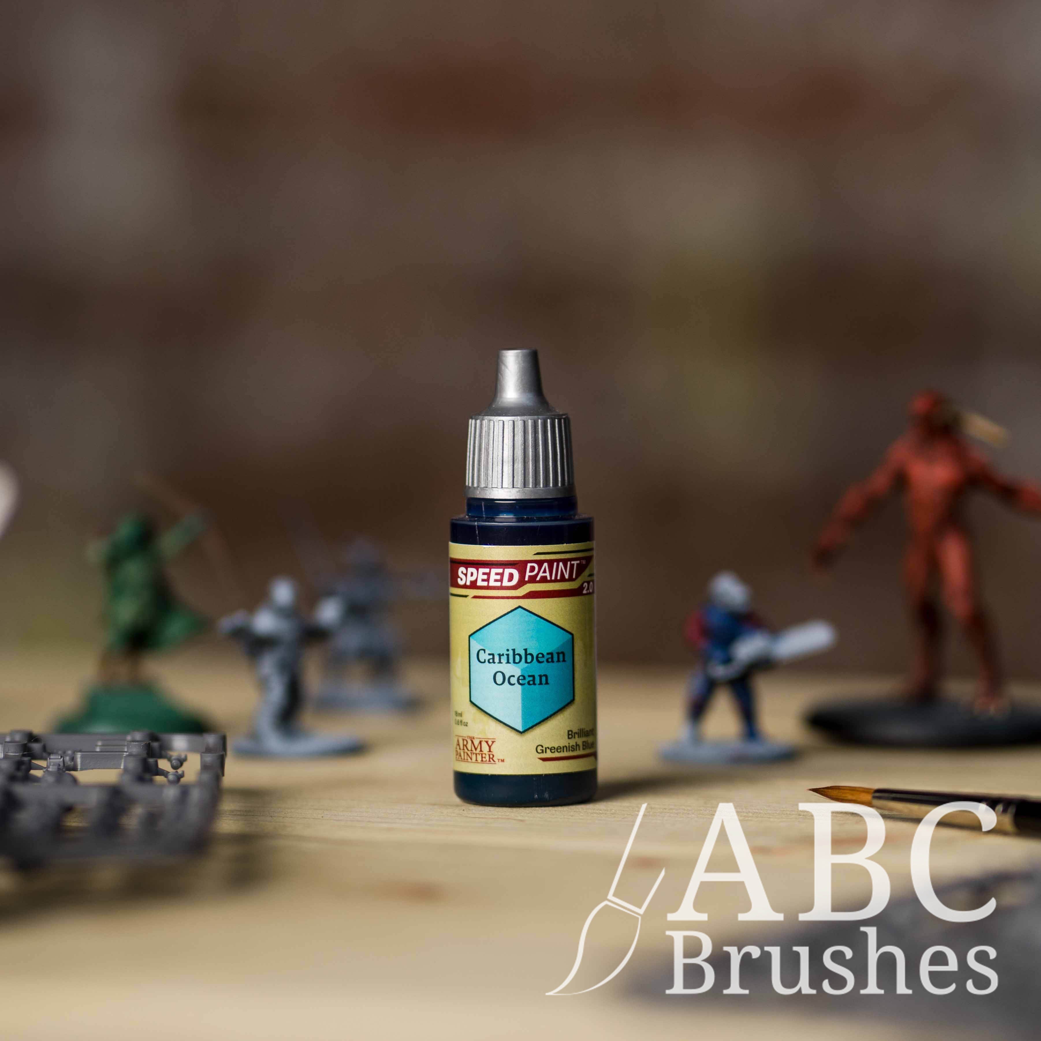 Bottle of the Army Painters Speedpaint in the colour Caribbean Ocean, with wargaming models and an ABC paintbrush in the background, on a wooden surface.