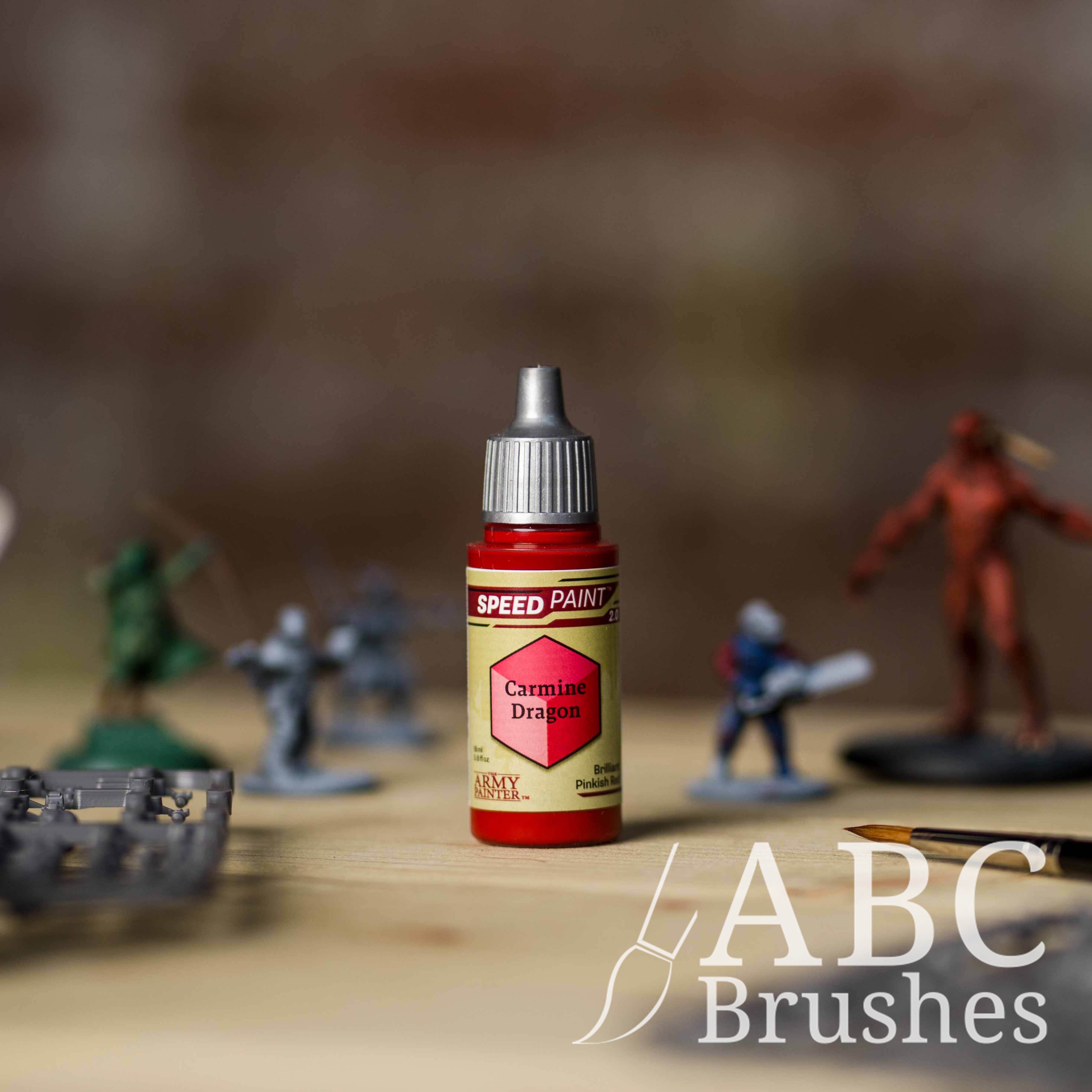 Bottle of the Army Painters Speedpaint in the colour Carmine Dragon, with wargaming models and an ABC paintbrush in the background, on a wooden surface.