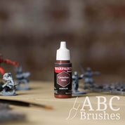 Bottle of the Army Painter Fanatics in the colour Carnelian Skin with various wargaming models and an ABC paintbrush in the background, on a wooden surface.