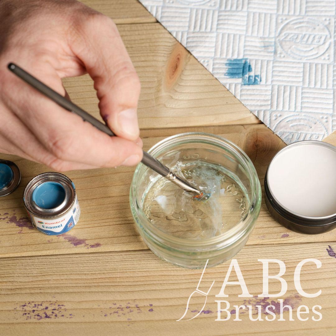 Cleaning an ABC paintbrush in mineral spirits, rinsing off any enamel paint on it. There is a Humbrol Enamels paint pot, ABC brush cleaner and kitchen towel next to this. 