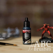 Bottle of the Army Painter Fanatics Metallic paint in the colour Cobalt Metal with various wargaming models and an ABC paintbrush in the background, on a wooden surface.