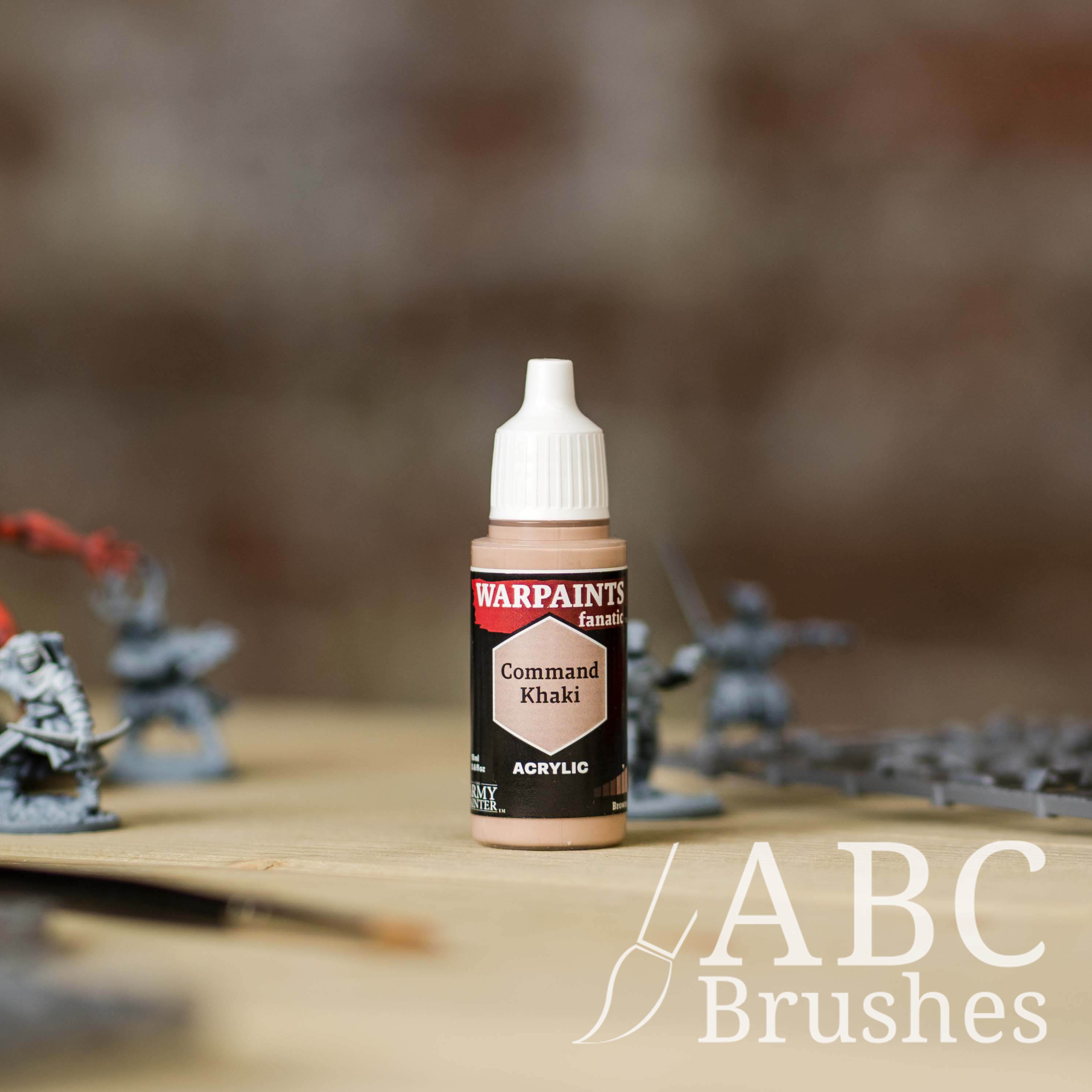 Bottle of the Army Painter Fanatics in the colour Command Khaki with various wargaming models and an ABC paintbrush in the background, on a wooden surface.